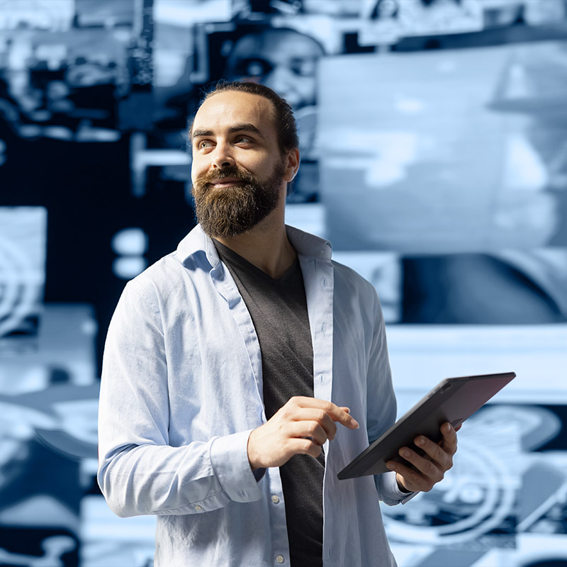 Un homme barbu qui se tient debout en souriant et qui utilise une tablette comme support.